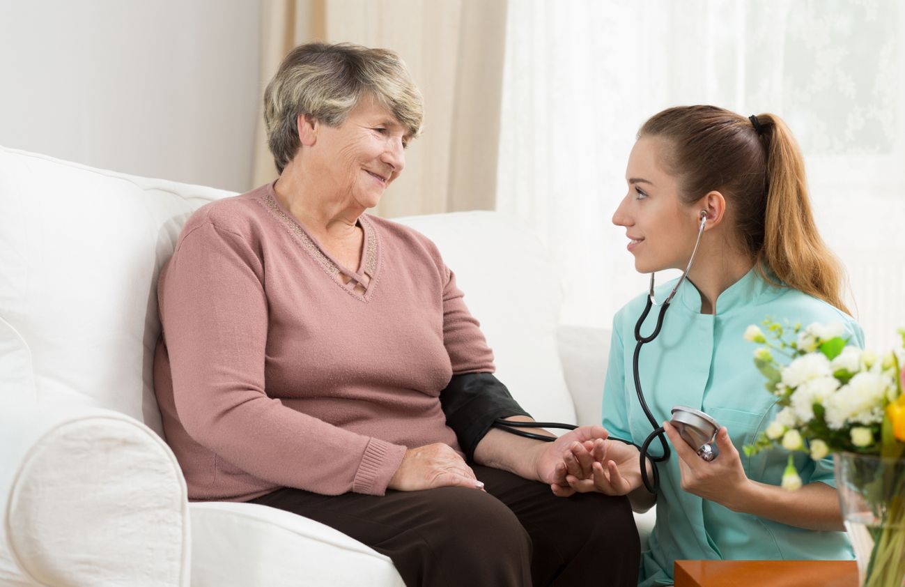 Eine Frau sitzt auf einer Couch, während eine Pflegerin ihren Puls misst.